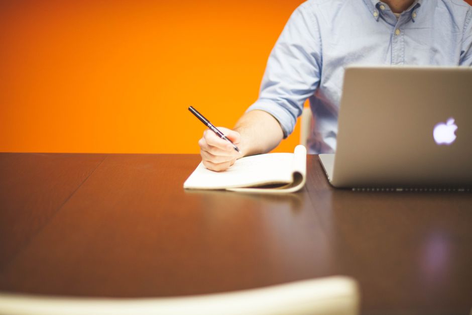 Persona escribiendo notas en un cuaderno junto a una laptop en un escritorio de madera